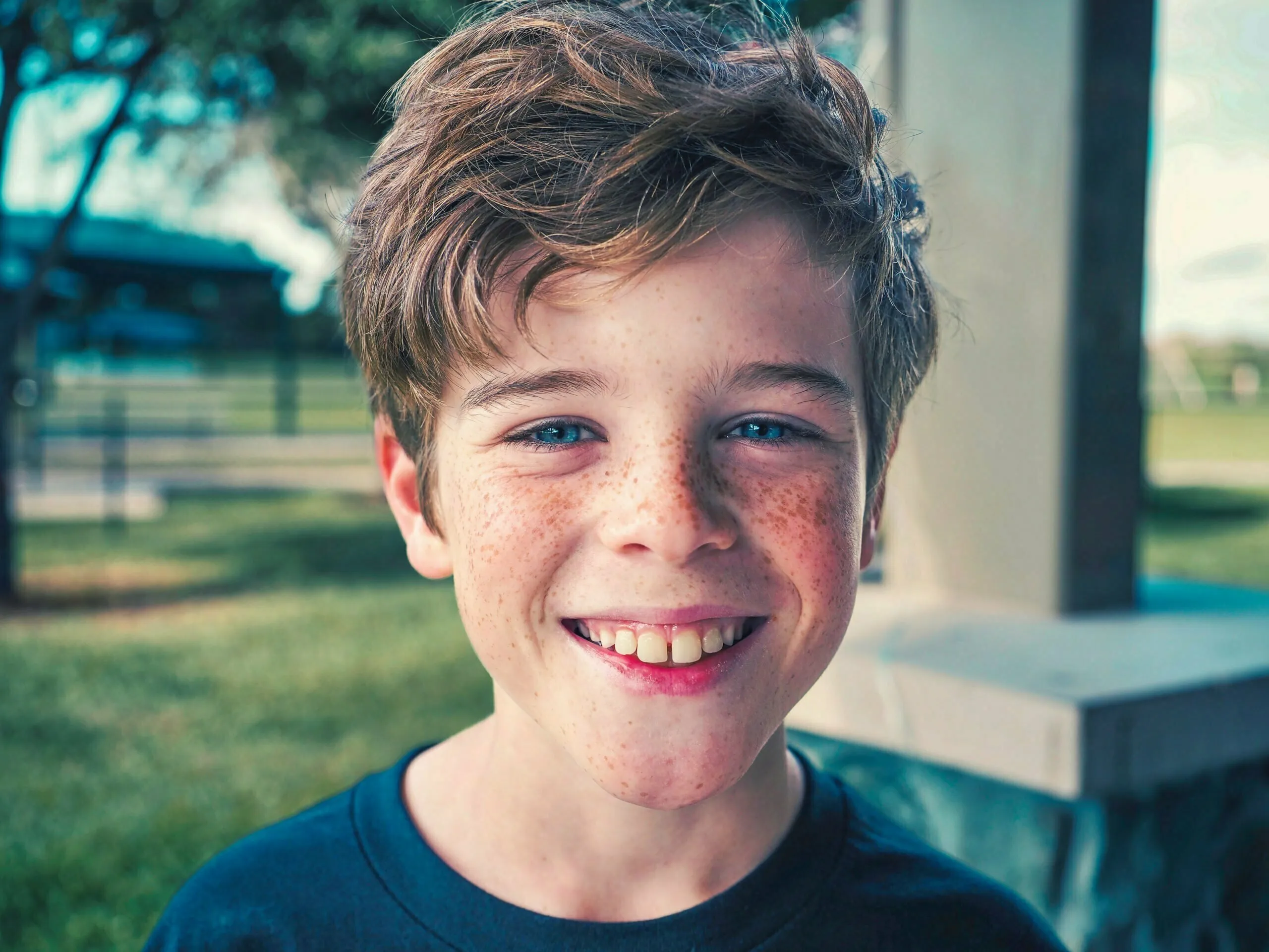Freckled boy around age 11 with brown hair grinning widely outdoors in a park setting