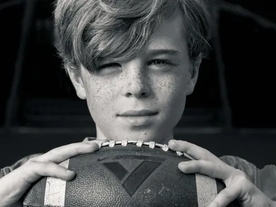 Black and white portrait of a freckled teen boy resting his chin on a football with a confident gaze