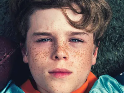 Freckled boy lying on turf grass next to a football, gazing upward in orange and teal jersey