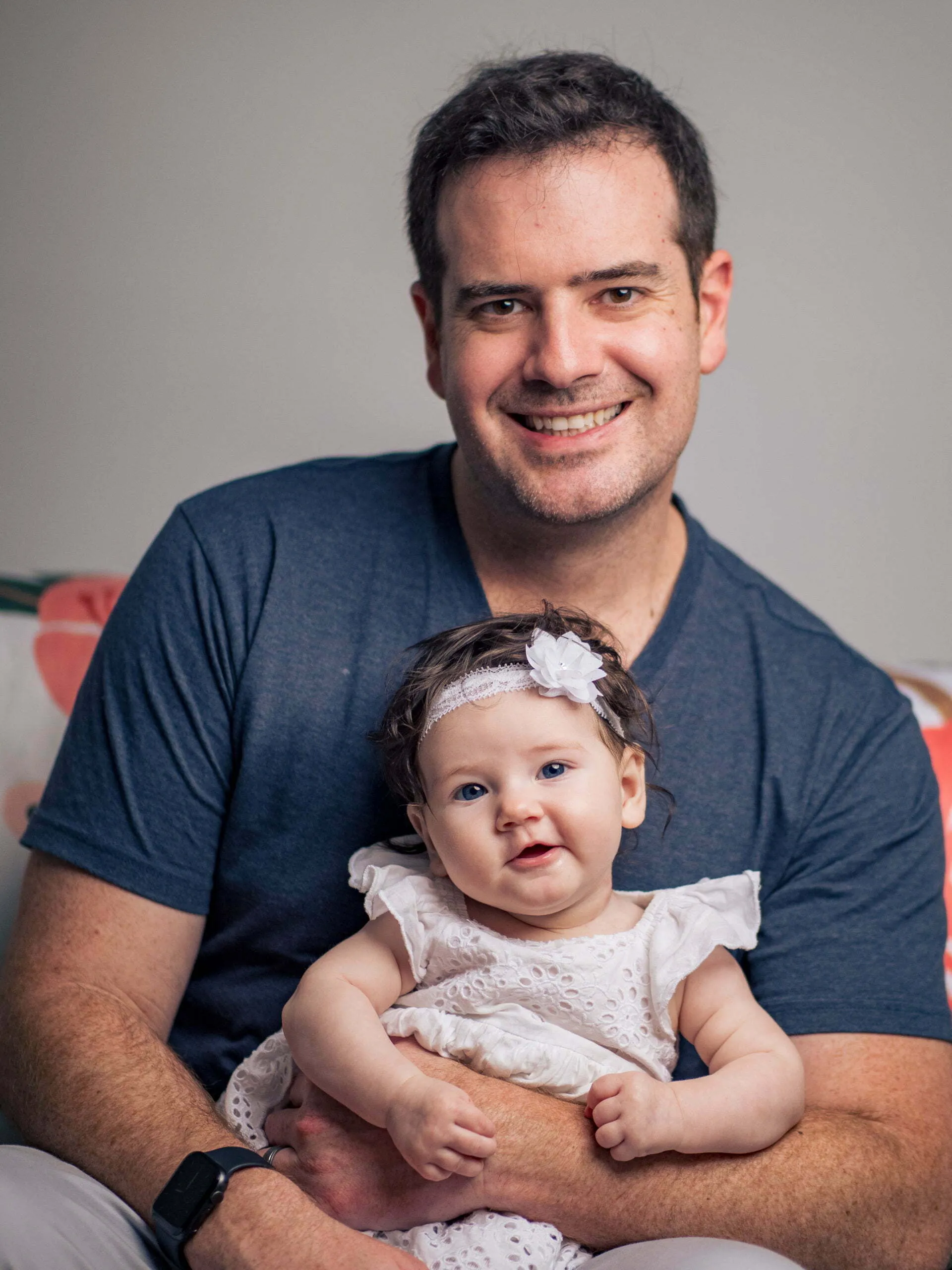 Smiling father in blue t-shirt holding infant daughter wearing white headband and lace dress indoors