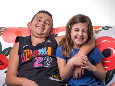 Brother and sister laughing together on a couch, boy in Miami basketball jersey and girl in blue top