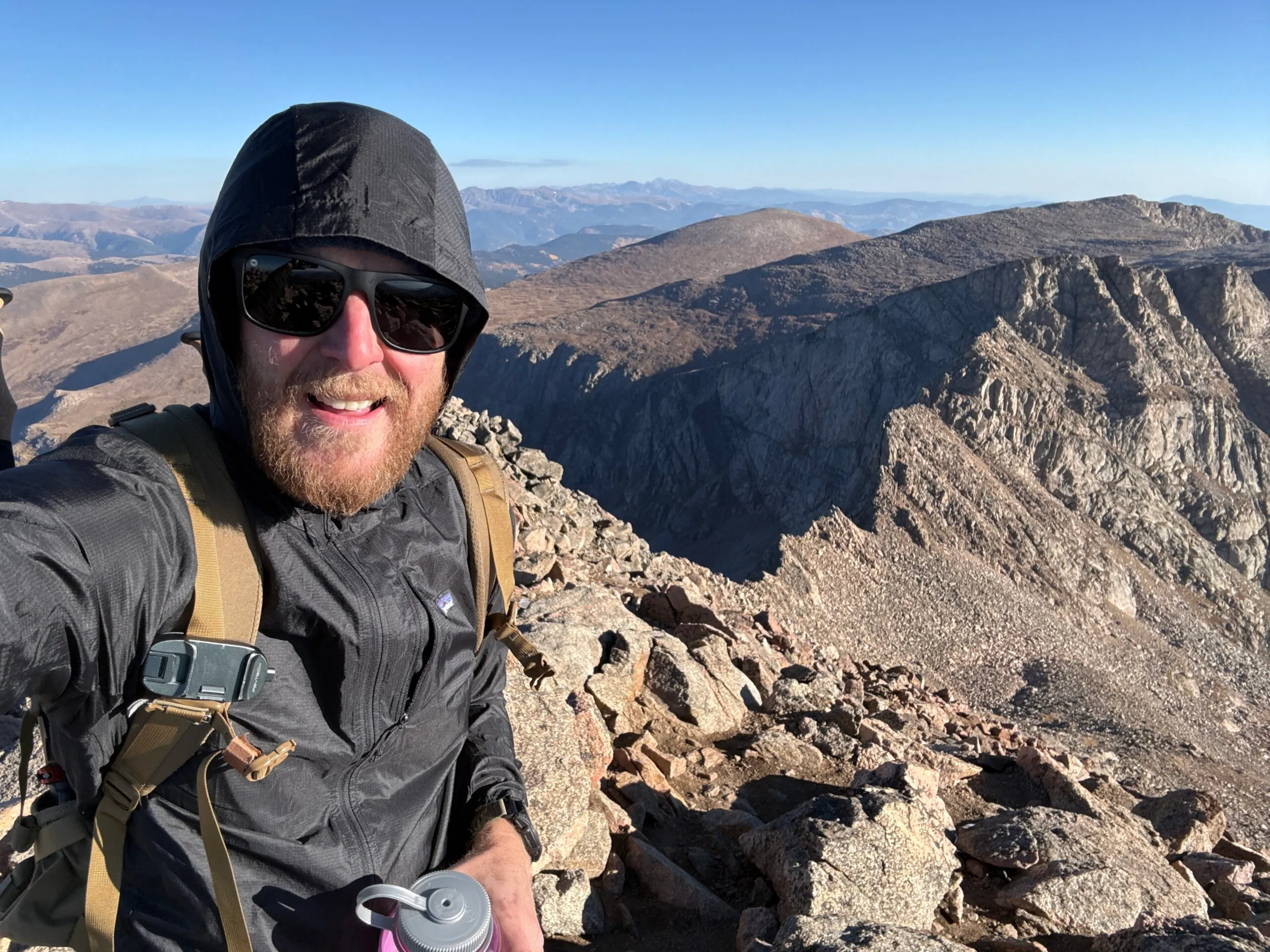 Bearded hiker in hooded jacket and sunglasses smiling on a rocky mountain summit with ridgelines stretching to the horizon