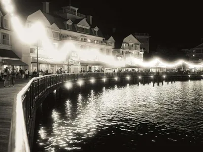 Black and white nighttime waterfront boardwalk with string lights reflecting on calm water and resort buildings beyond