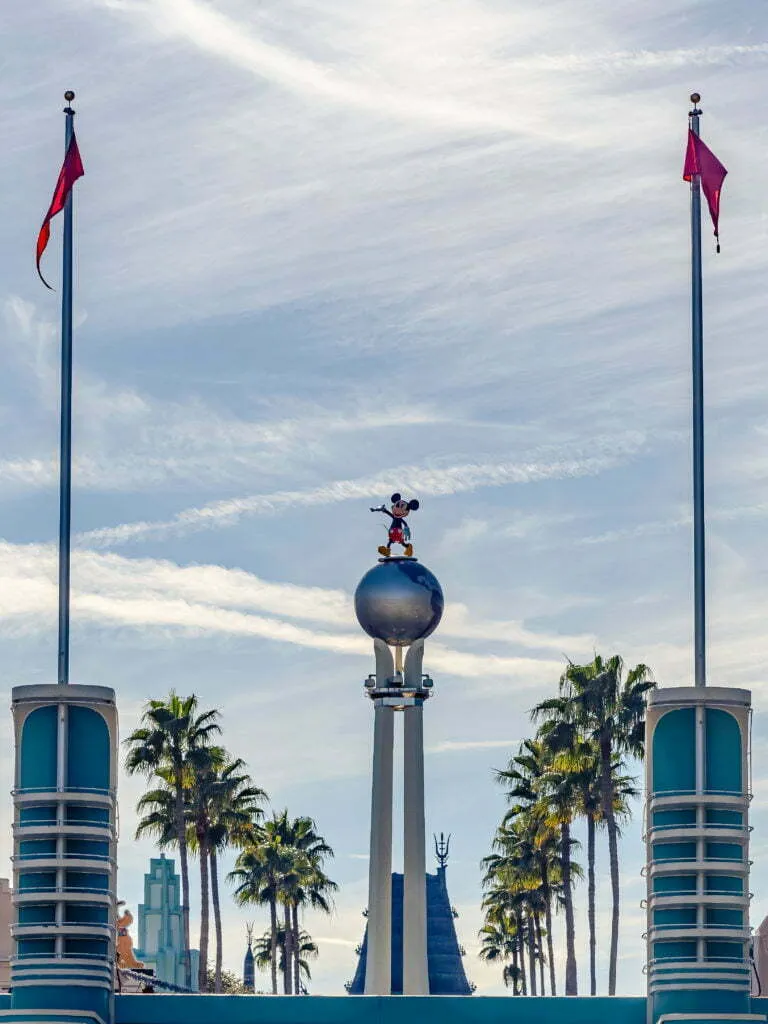 Theme park entrance with character figure atop a globe between two flags and palm trees under wispy clouds