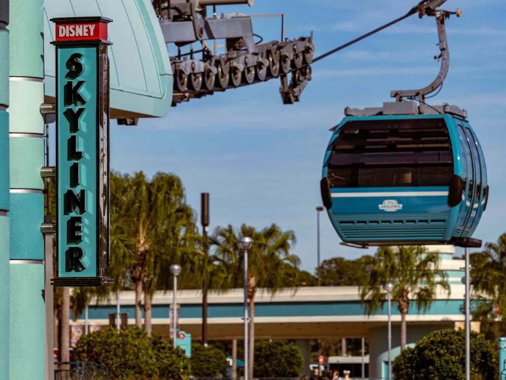 Teal gondola cabin departing a station with a neon Skyliner sign, palm trees, and blue sky in the background