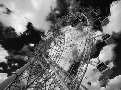 Black and white low-angle view of a giant observation wheel with enclosed gondolas against dramatic cloudy sky
