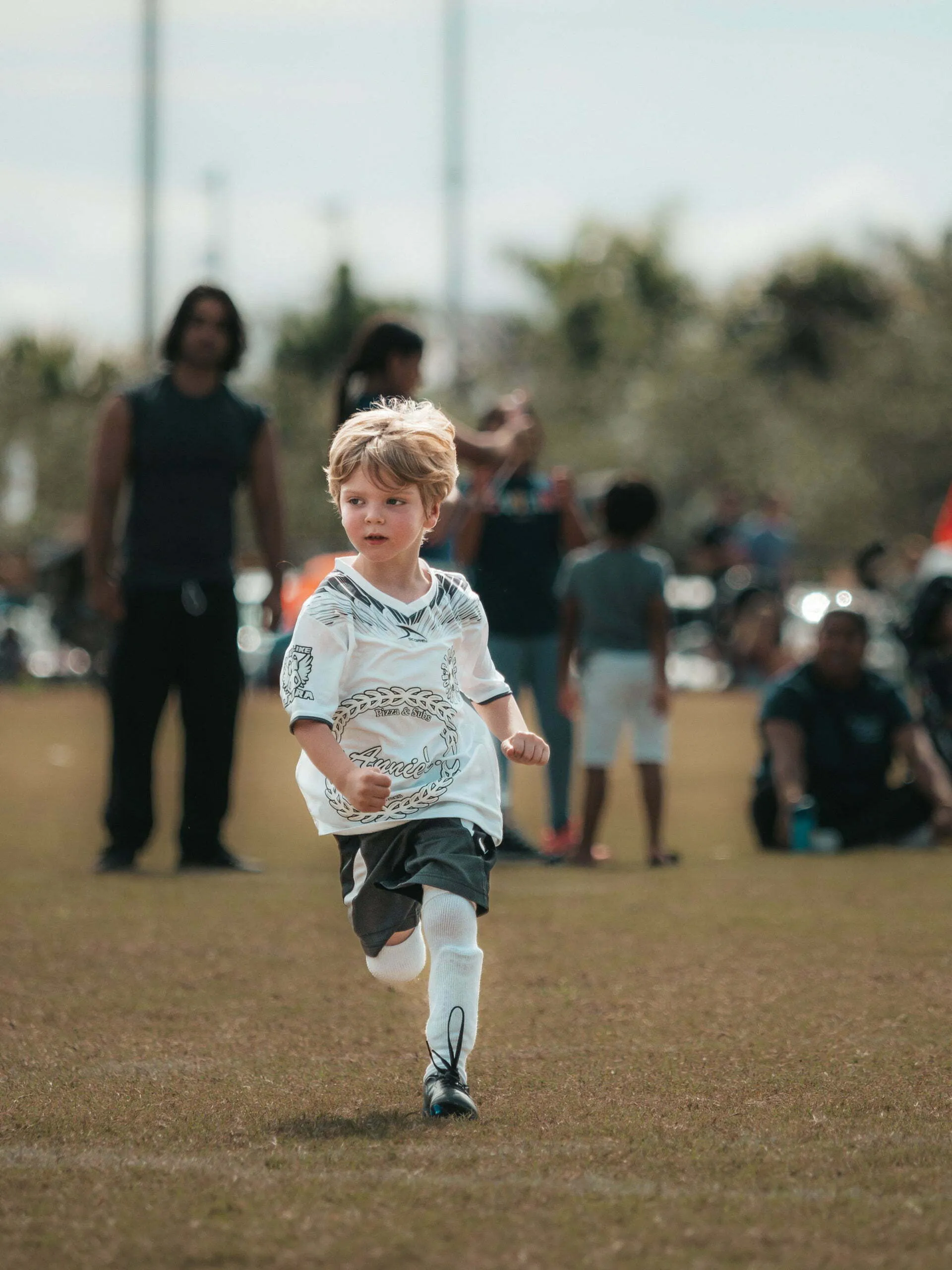 Young boy around age 5 sprinting across a soccer field in a white jersey and knee-high socks with spectators behind him