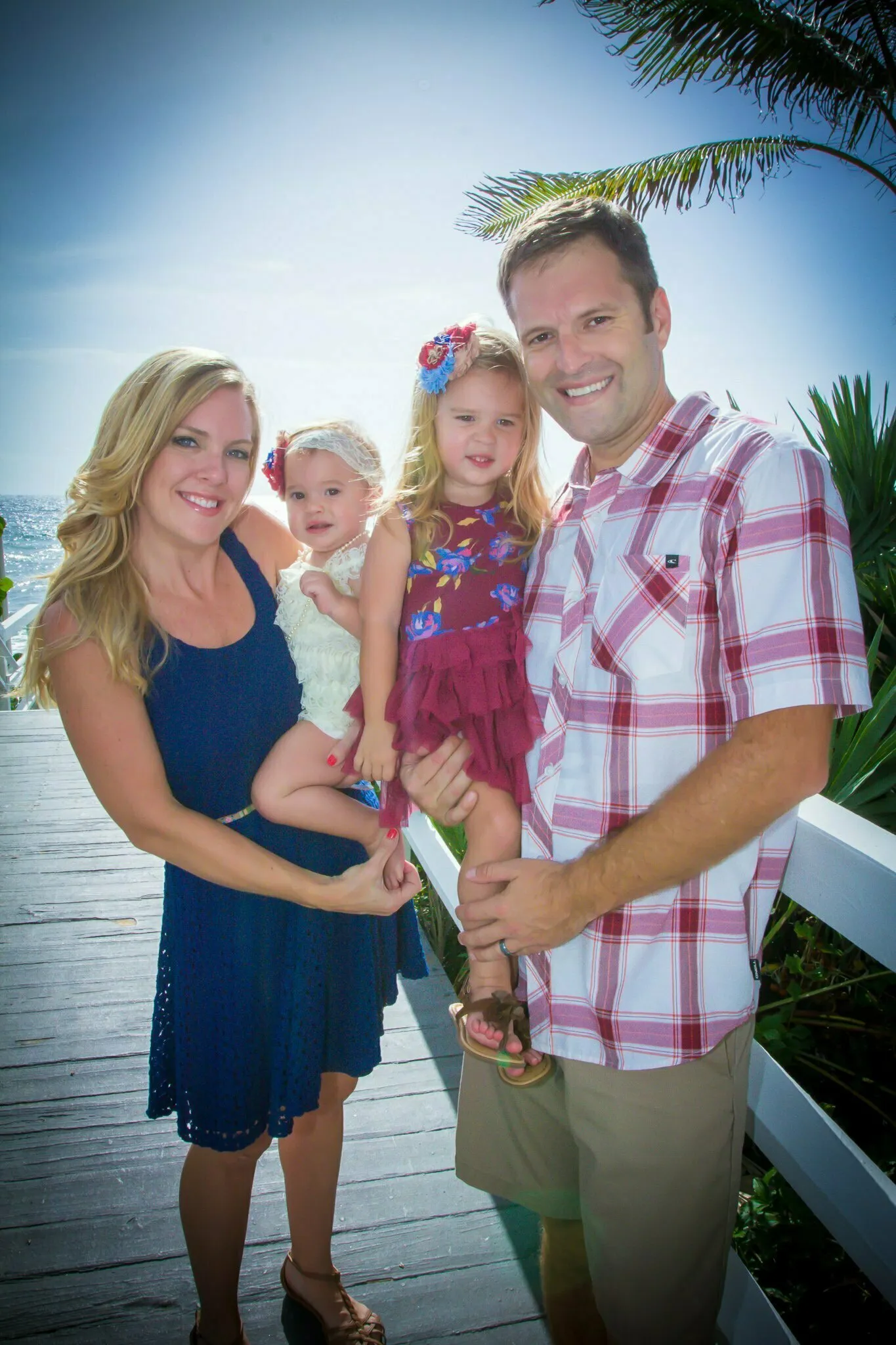 Smiling family of four with parents holding two young daughters on a sunny boardwalk with tropical plants behind them