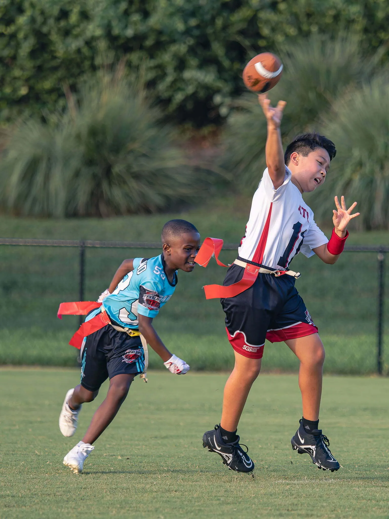 Two boys playing youth flag football, one throwing the ball while the other rushes to pull a flag on a green field