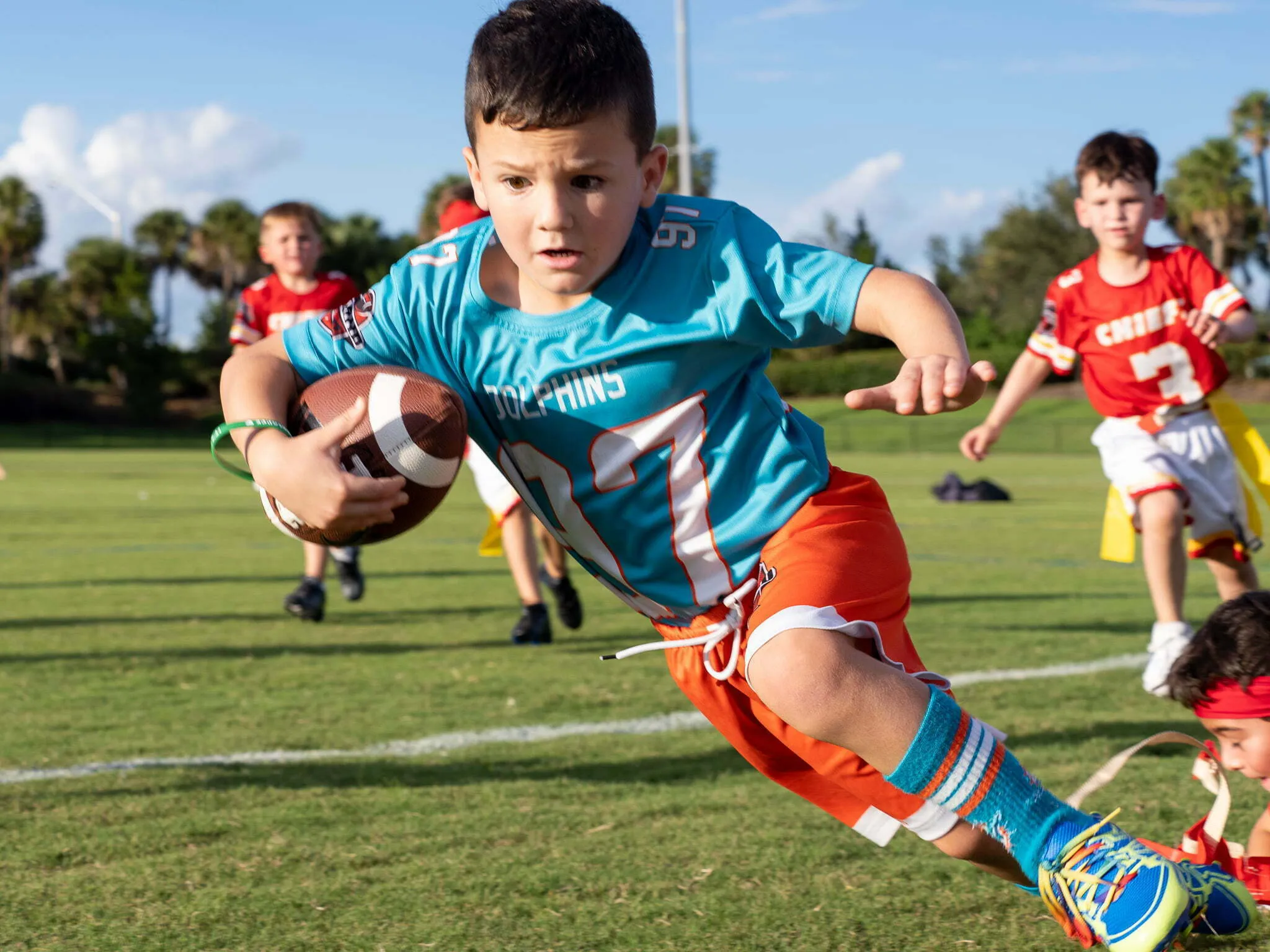 Young boy in teal Dolphins jersey diving forward with the football during a youth flag football game on a sunny day