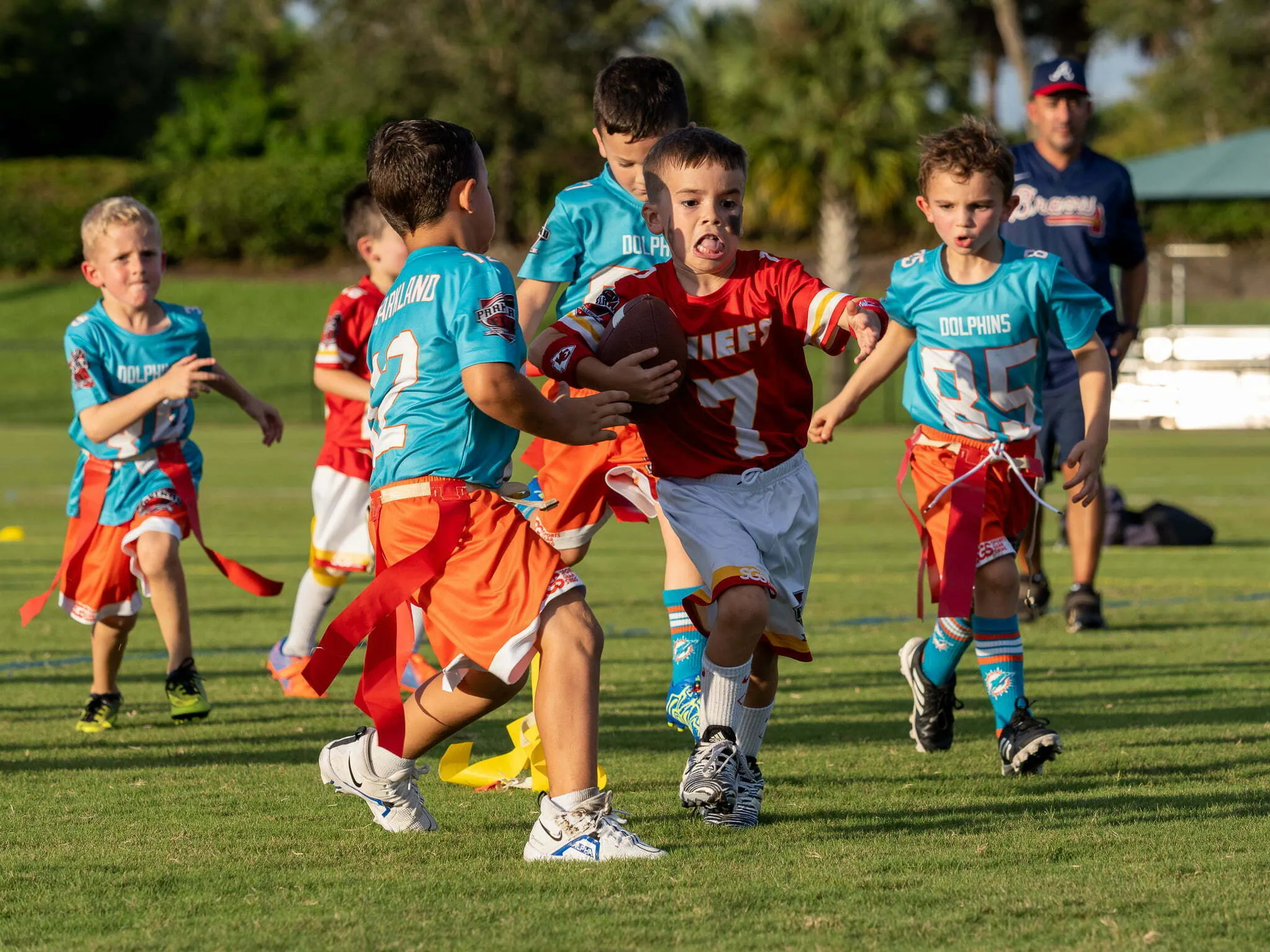 Boy in red Chiefs jersey running with the football through a crowd of Dolphins defenders during youth flag football
