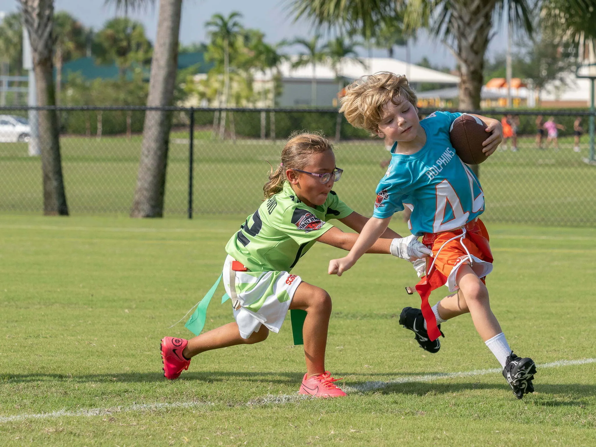 Girl in green jersey reaching to pull the flag of a boy in teal Dolphins jersey carrying the football on a sunny field