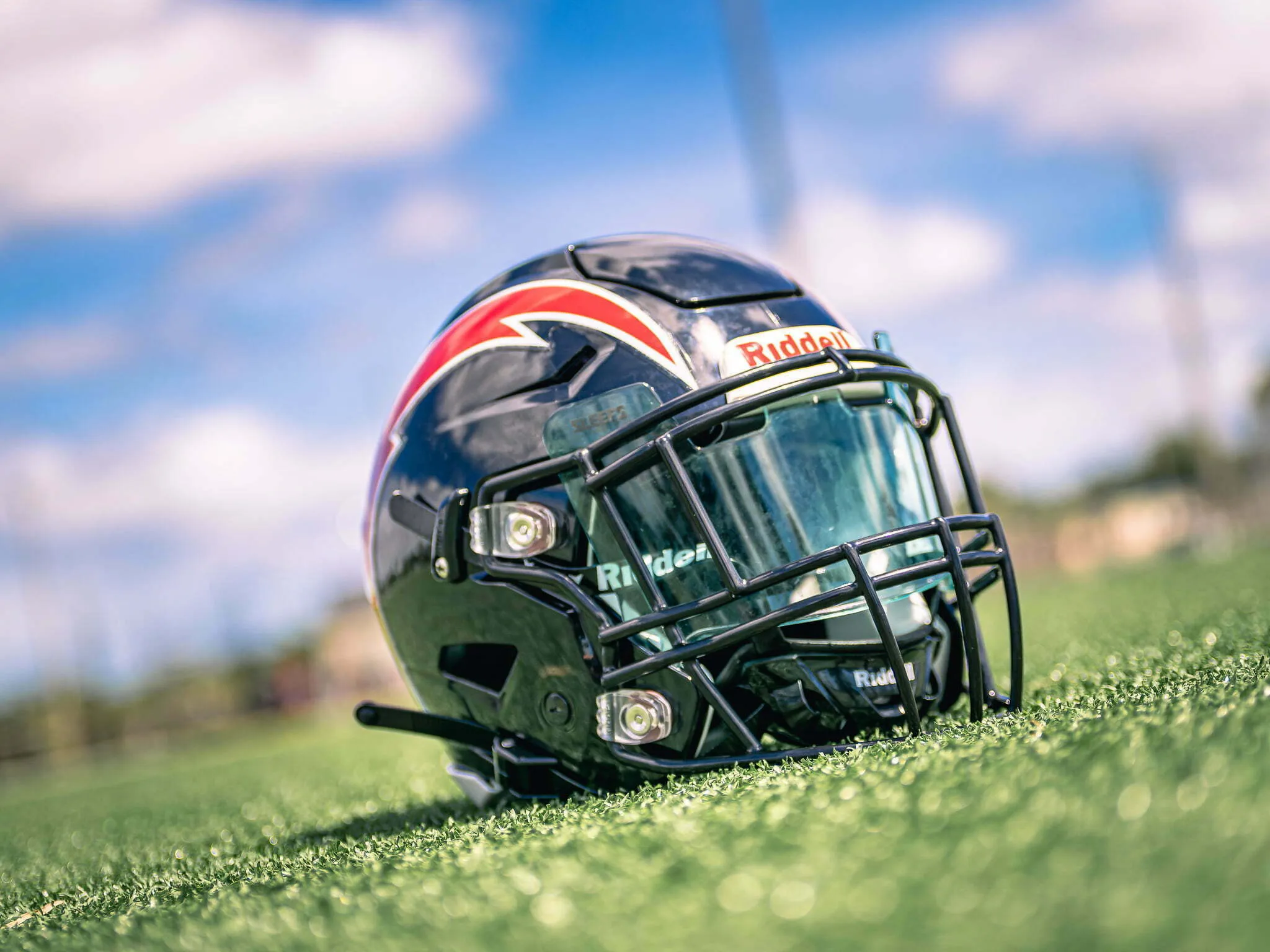 Dark blue Riddell football helmet with red stripe resting on green turf under a blue sky with white clouds