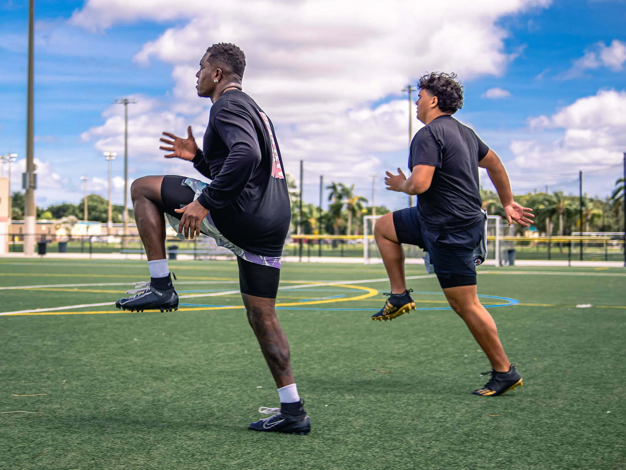 Two football players in black shirts warming up with high knee stretches on a turf field under a blue sky