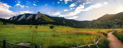 Panoramic view of Boulder Flatirons with a dirt trail, wooden fence, green meadow, and golden grasses at sunset