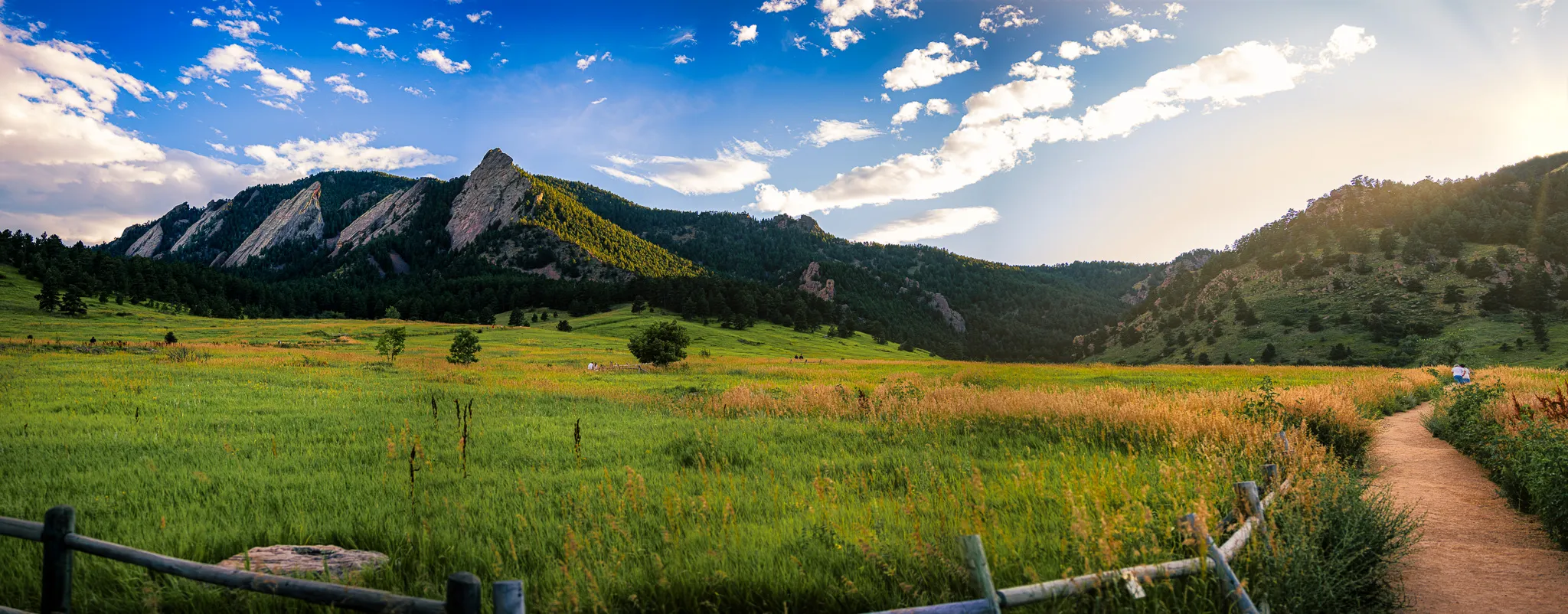Panoramic view of Boulder Flatirons with a dirt trail, wooden fence, green meadow, and golden grasses at sunset