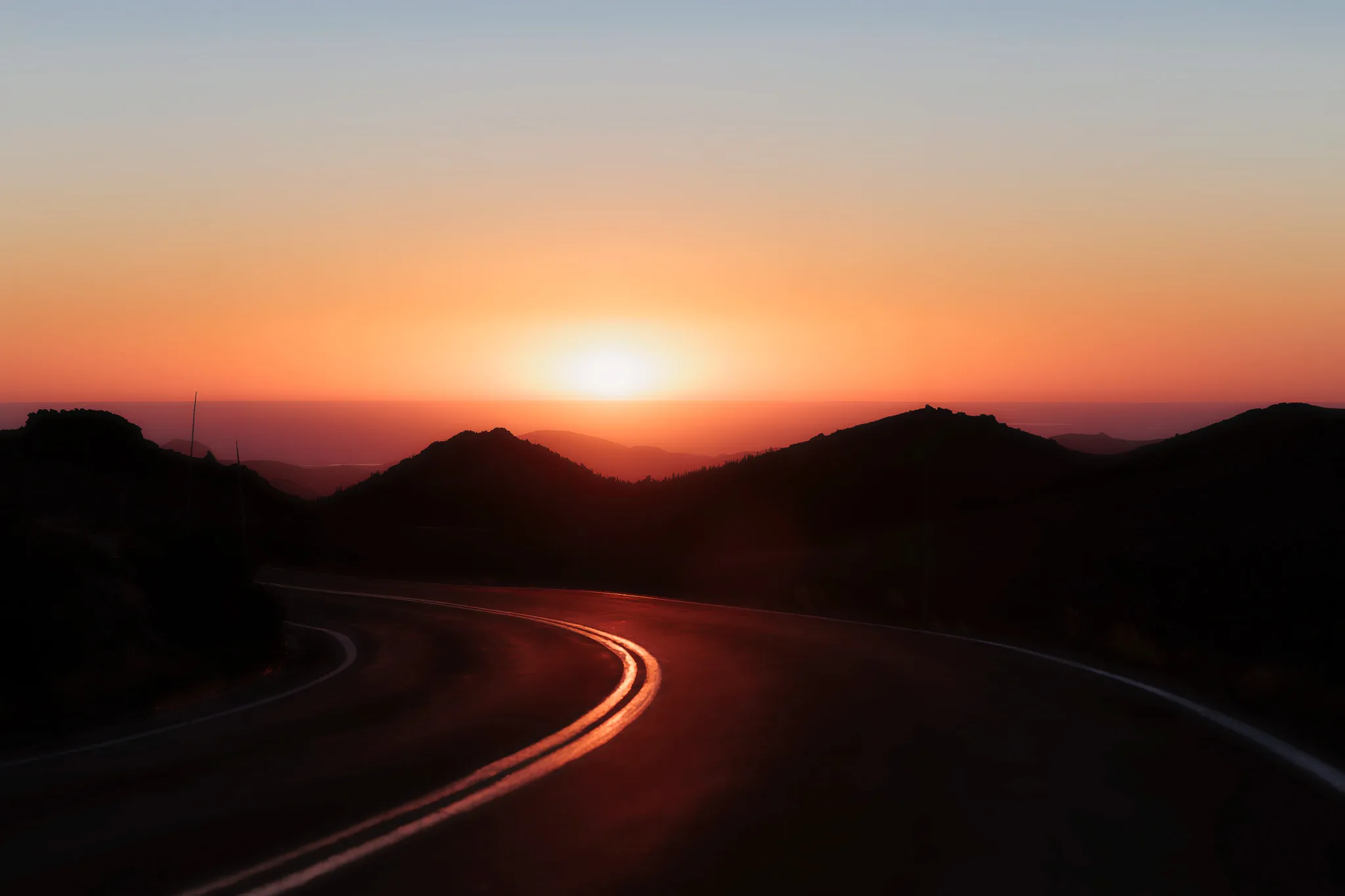 Curving mountain road silhouetted against a golden orange sunset with hills in the distance