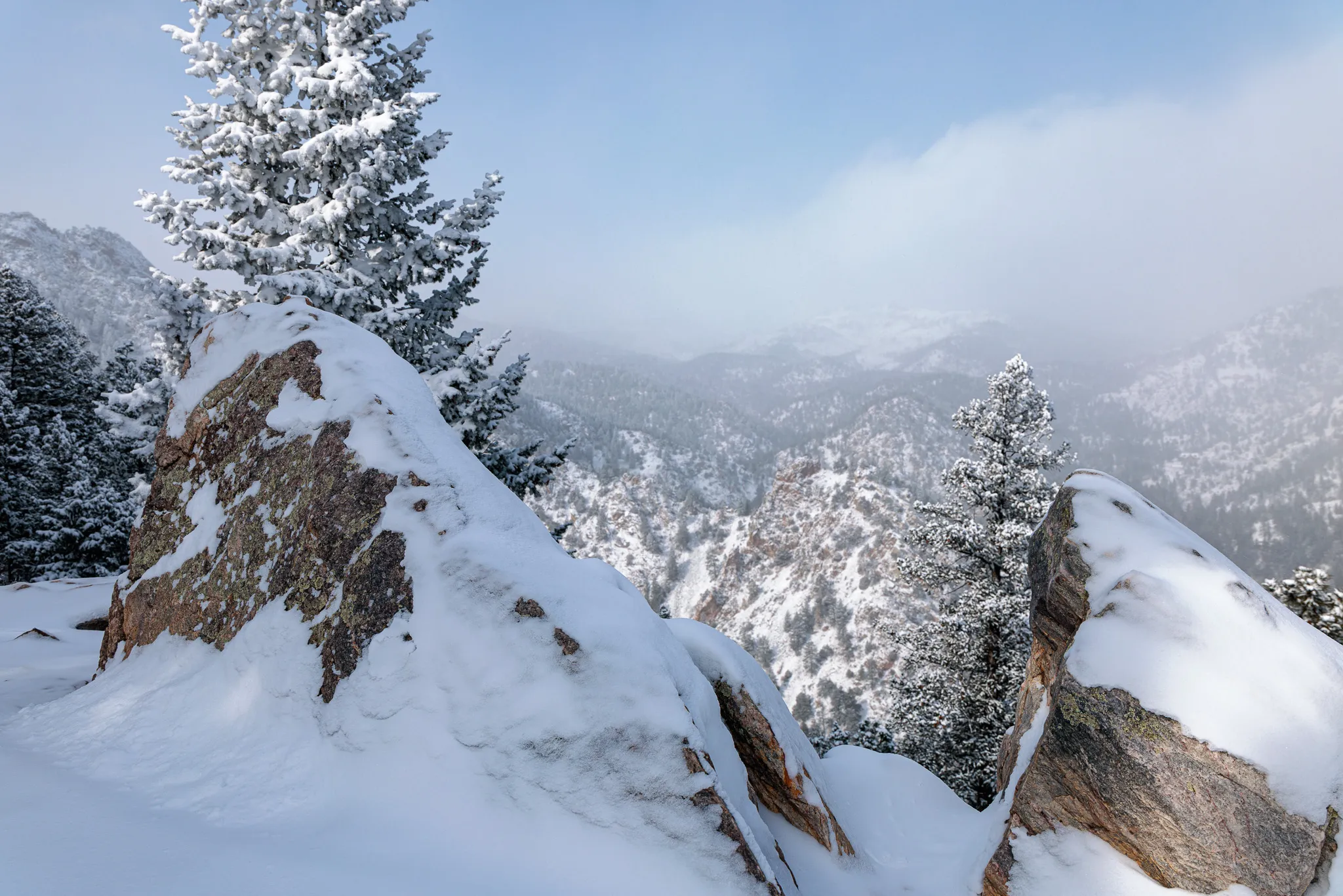 Snow-blanketed rock outcrop overlooking a misty mountain valley with frosted evergreens on an overcast day