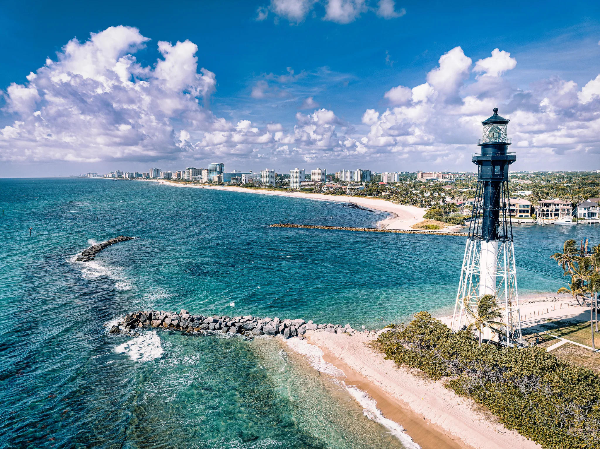 Aerial drone view of a coastal lighthouse on a sandy point with turquoise ocean water, beach, and city skyline under bright clouds