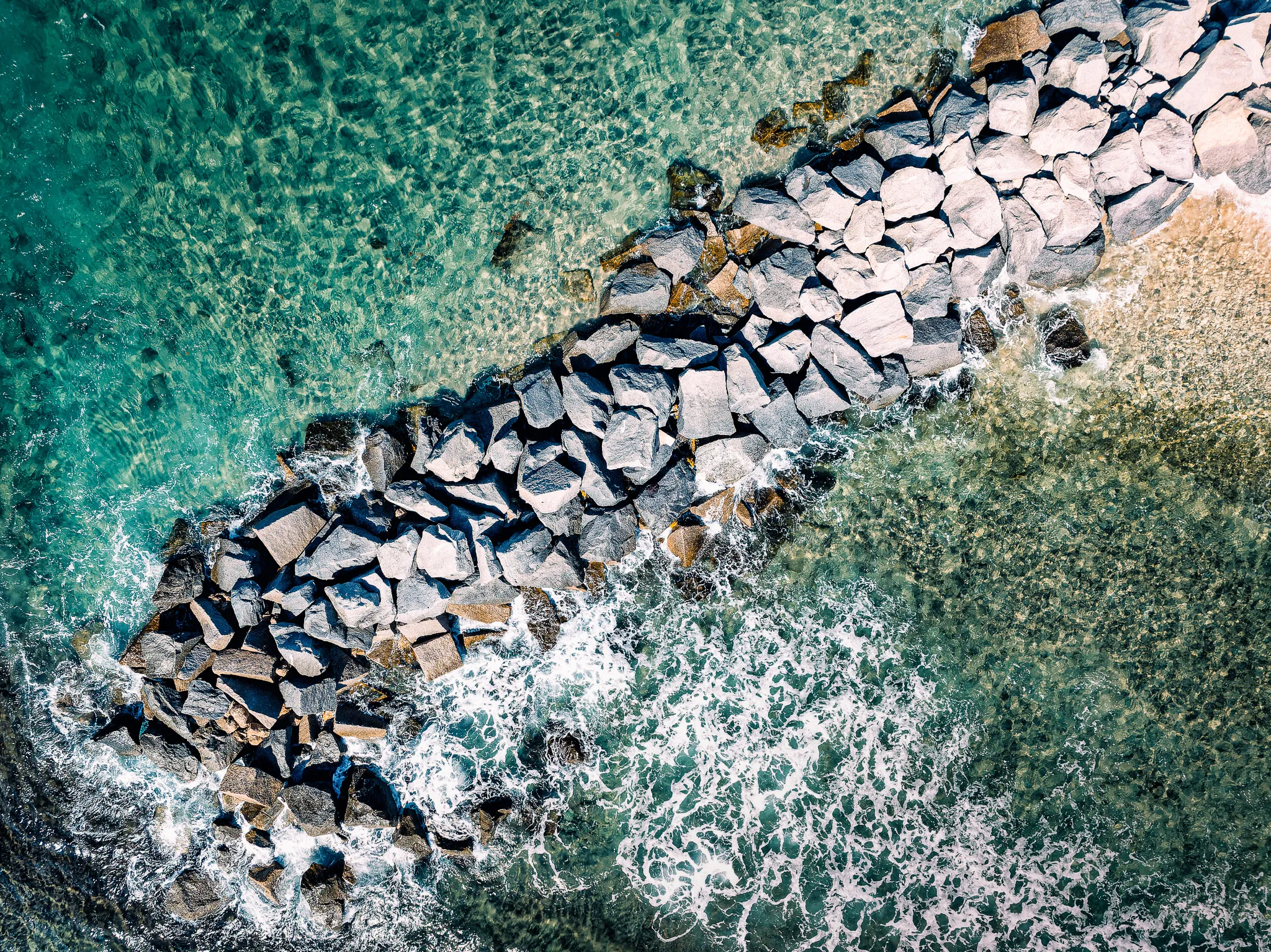 Top-down aerial view of a rock jetty cutting through turquoise ocean water with waves breaking against the stones