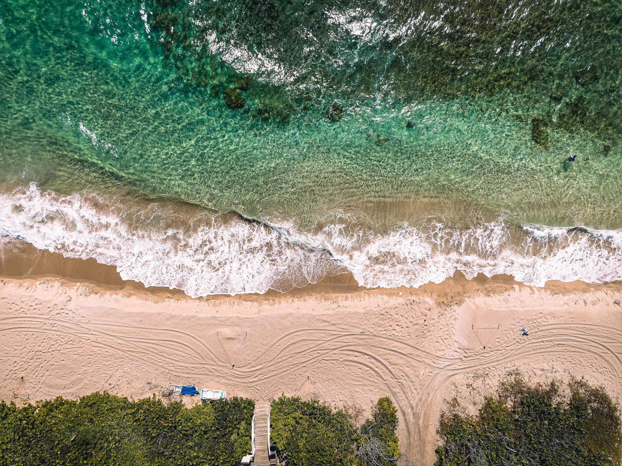 Aerial drone view of turquoise ocean waves breaking onto sandy beach with tropical tree canopy on both sides