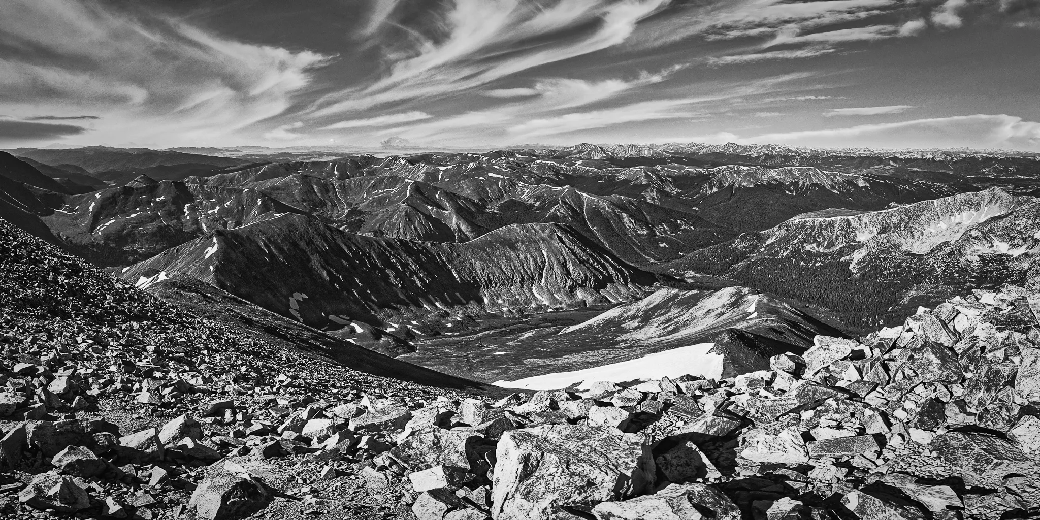Black and white panoramic view from a high Colorado summit with rocky foreground, alpine tundra, and dramatic wispy clouds