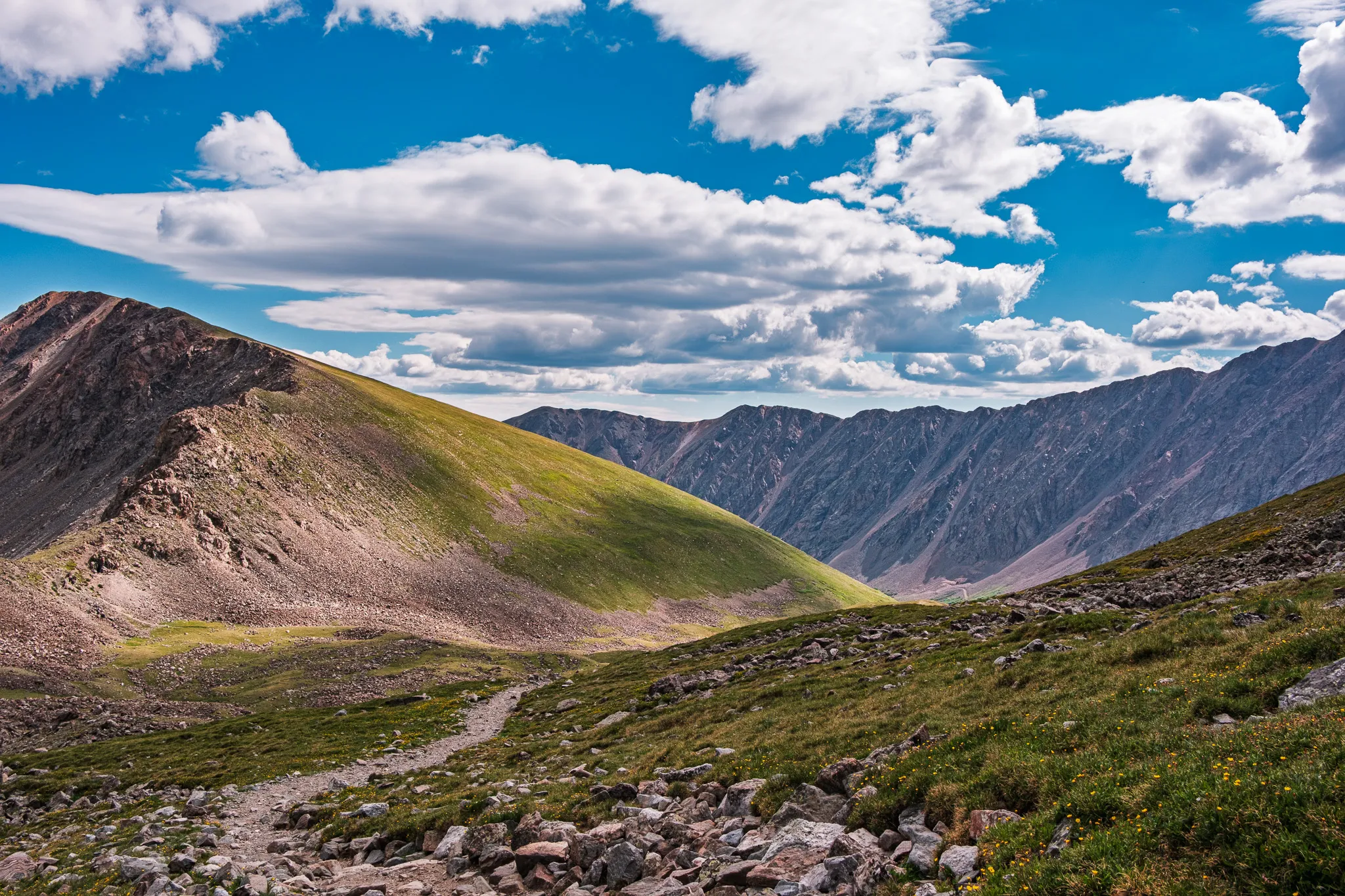 Alpine hiking trail winding through green tundra toward a high Colorado mountain ridge under puffy white clouds