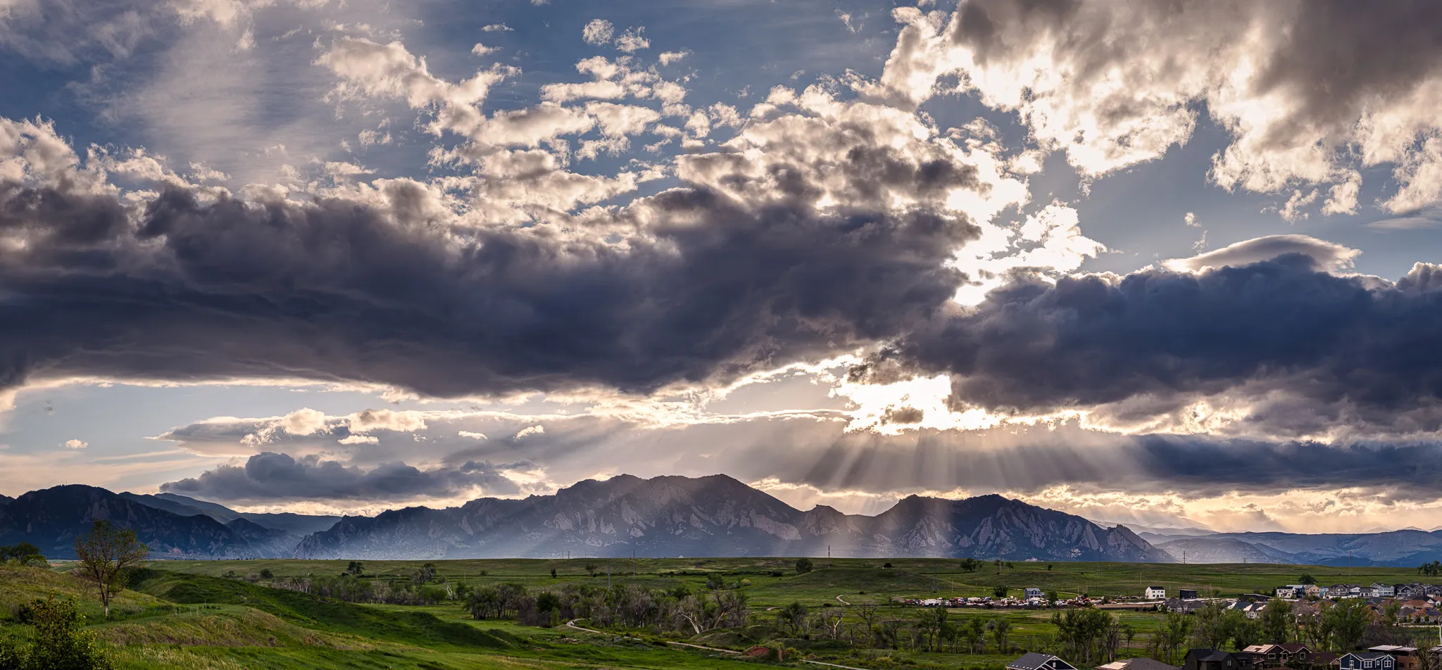 Panoramic view of Boulder Flatirons with dramatic sunbeams breaking through storm clouds over green foothills and valley