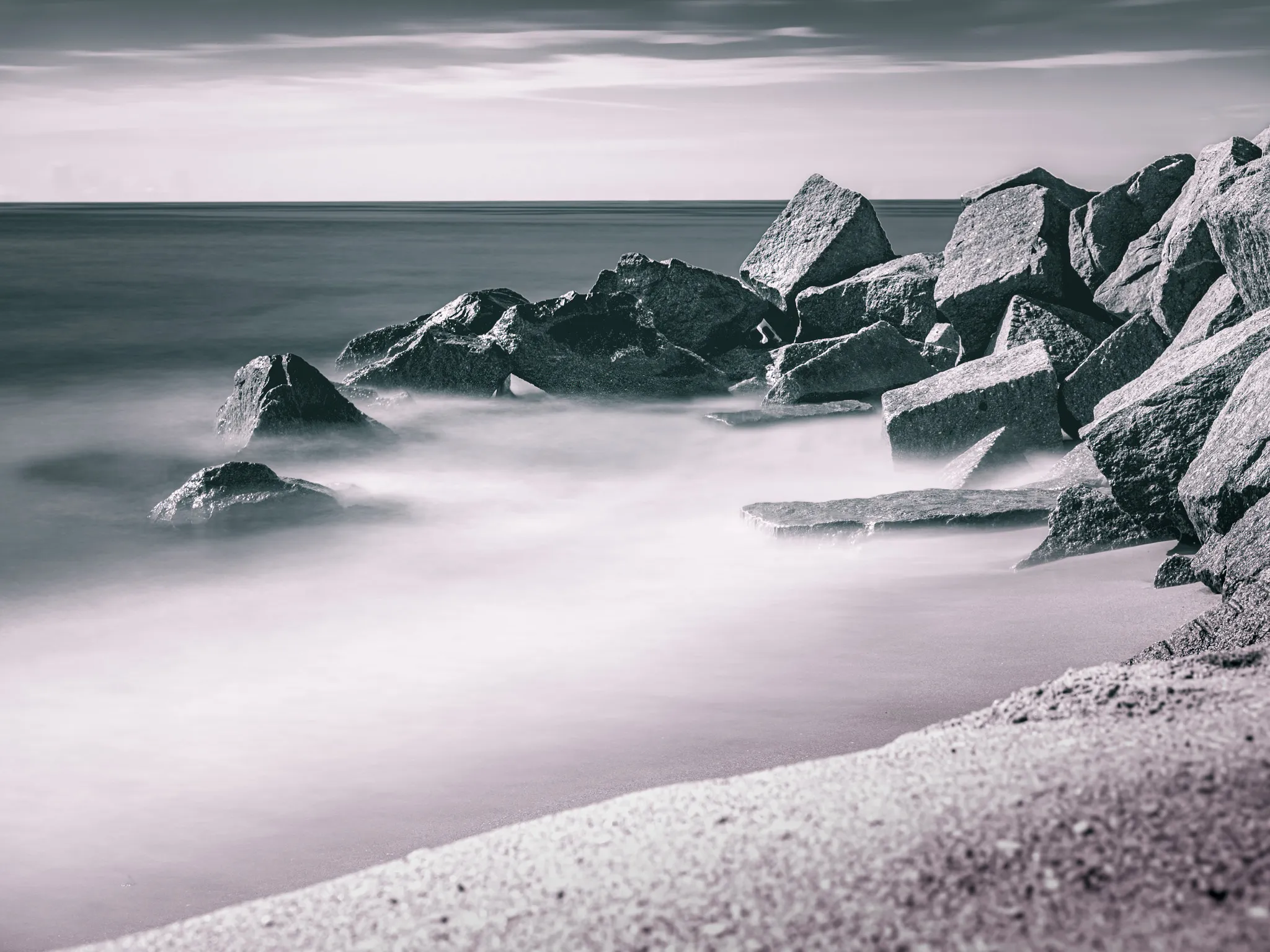 Black and white long exposure of large rocks on a sandy beach with smooth misty ocean water and overcast sky