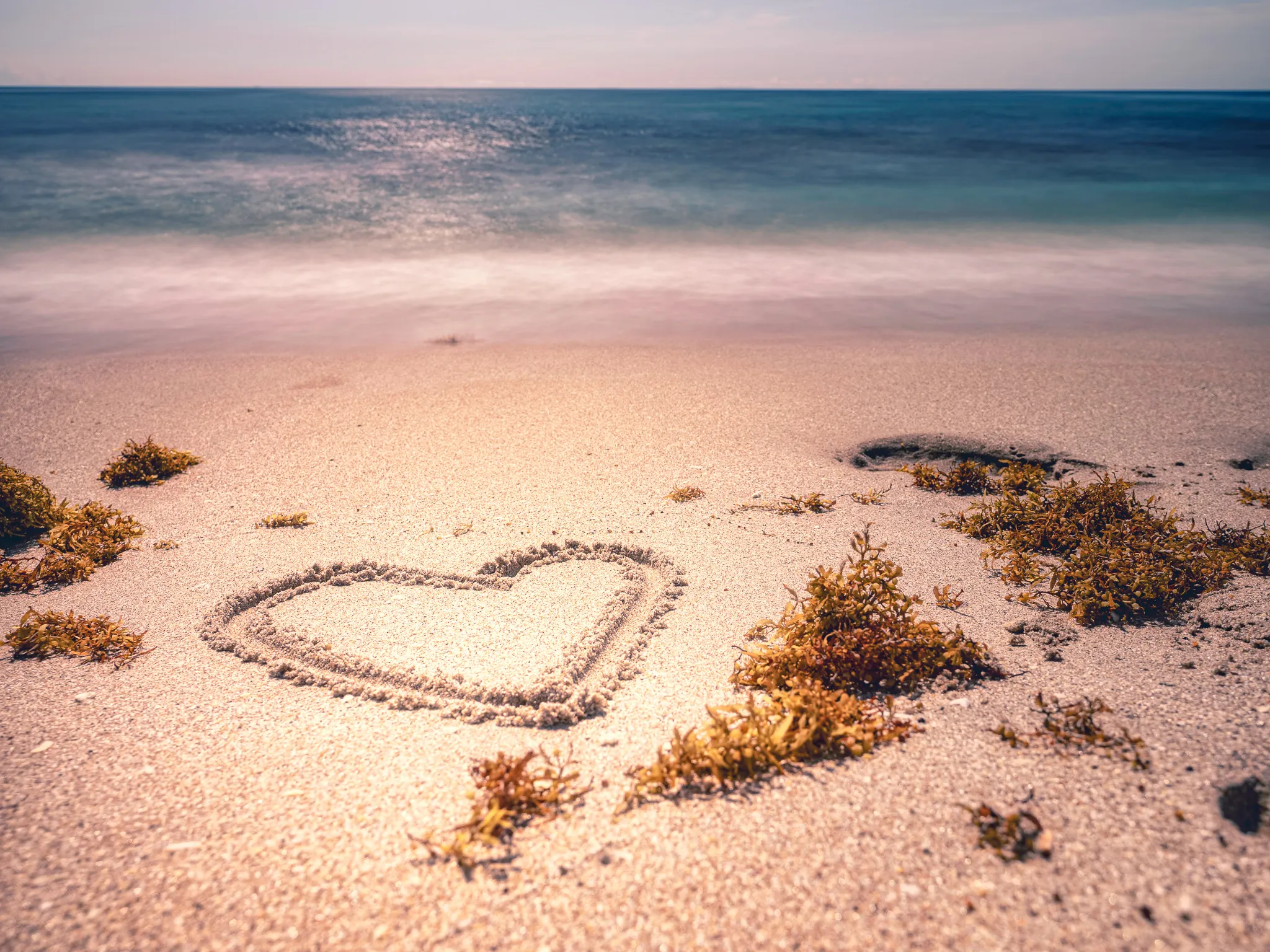 Heart shape drawn in sand on a tropical beach with turquoise ocean water, scattered seaweed, and clear sky