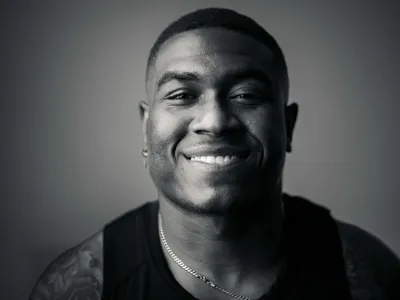 Close-up studio headshot of smiling young man with short hair, chain necklace, and tattoo, black and white