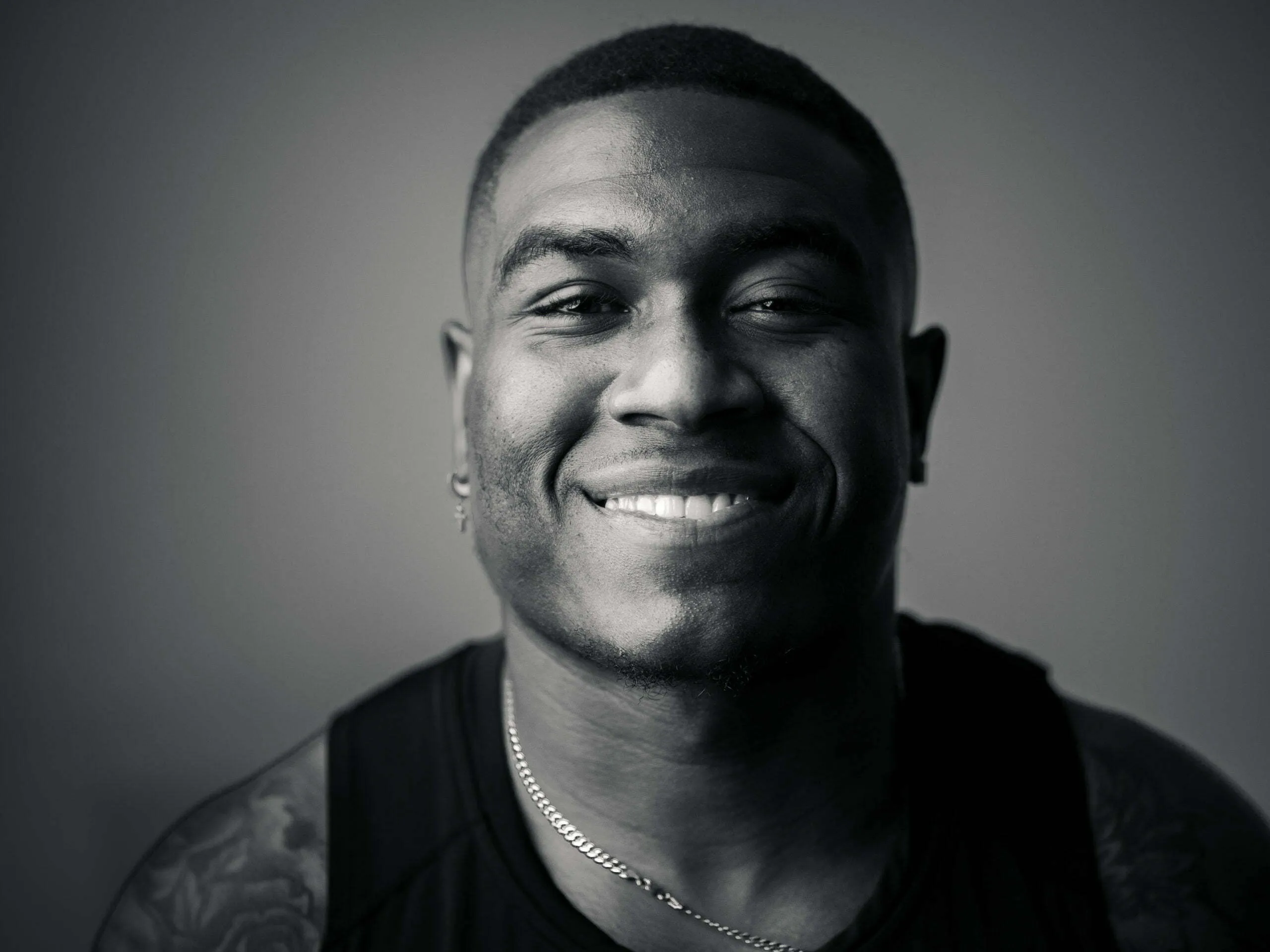 Close-up studio headshot of smiling young man with short hair, chain necklace, and tattoo, black and white