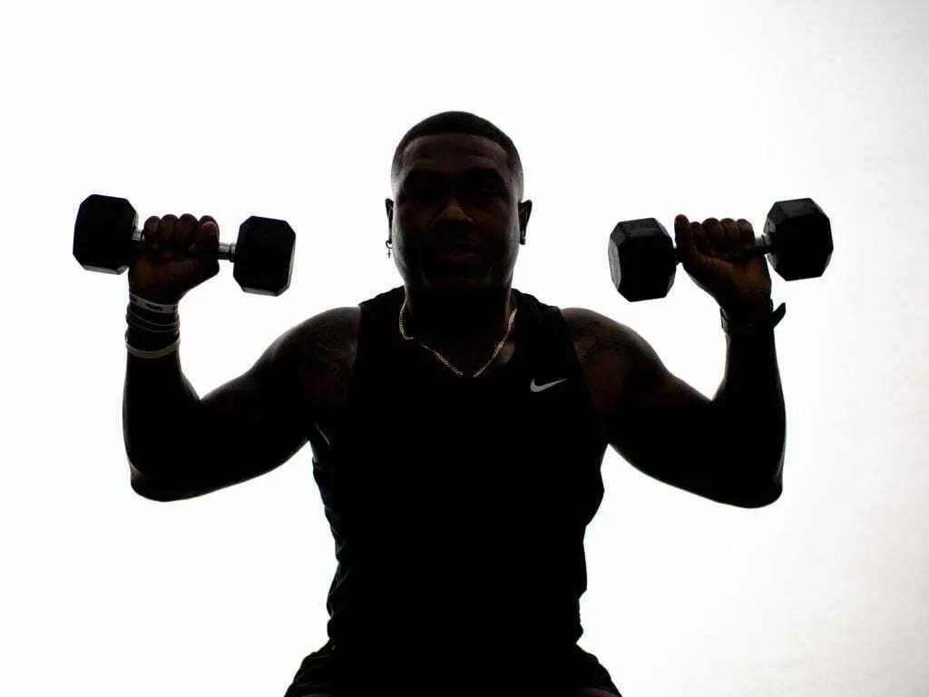 Athlete holding two dumbbells at shoulder height, backlit silhouette in Nike tank top, studio fitness portrait