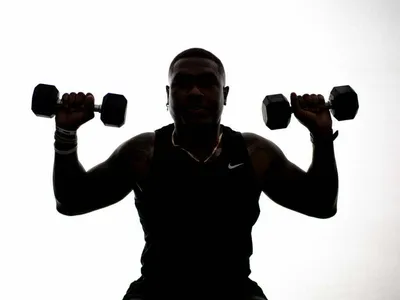 Athlete holding two dumbbells at shoulder height, backlit silhouette in Nike tank top, studio fitness portrait