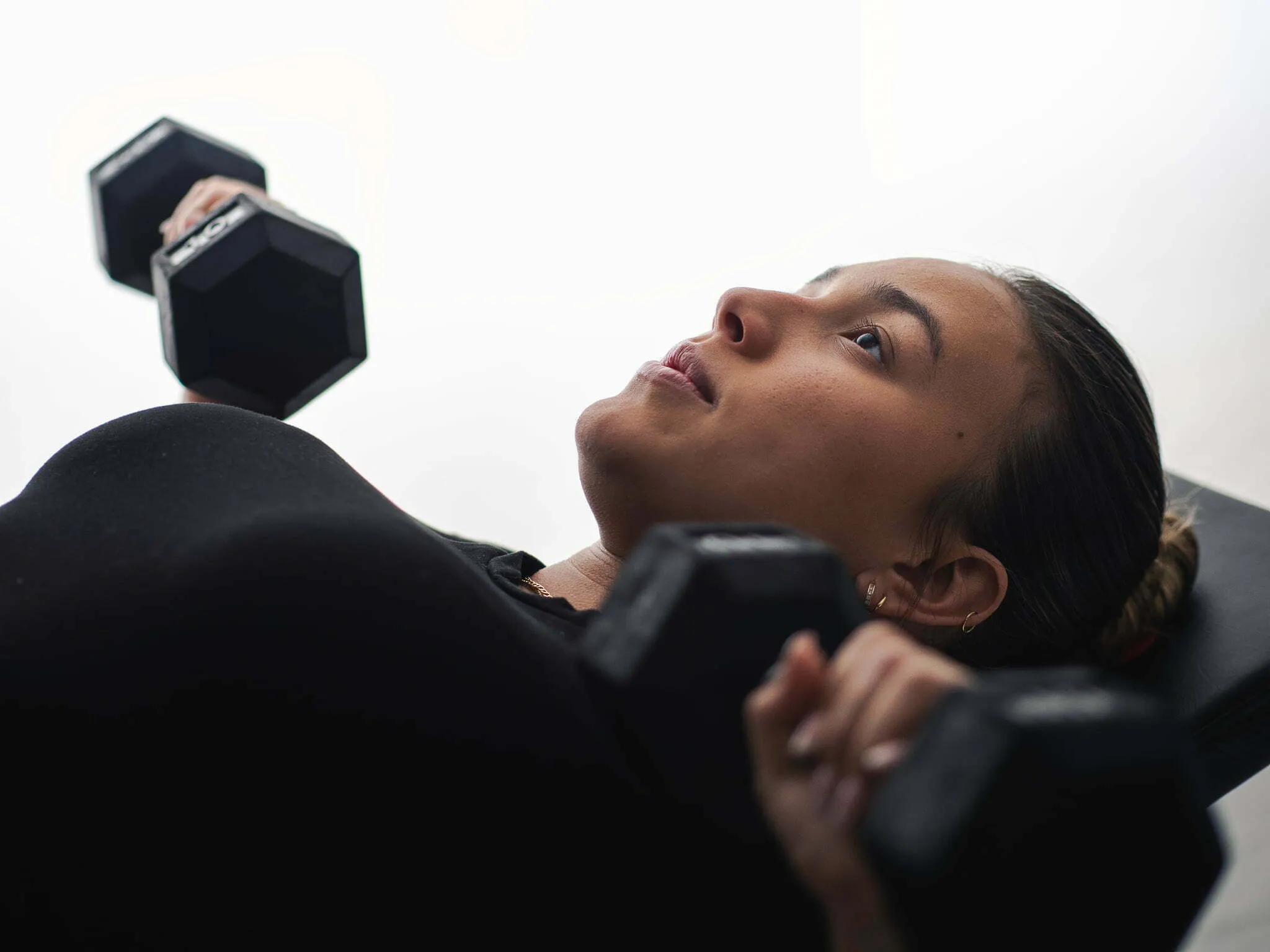 Young woman lying on weight bench pressing dumbbells overhead, focused expression, close-up fitness portrait
