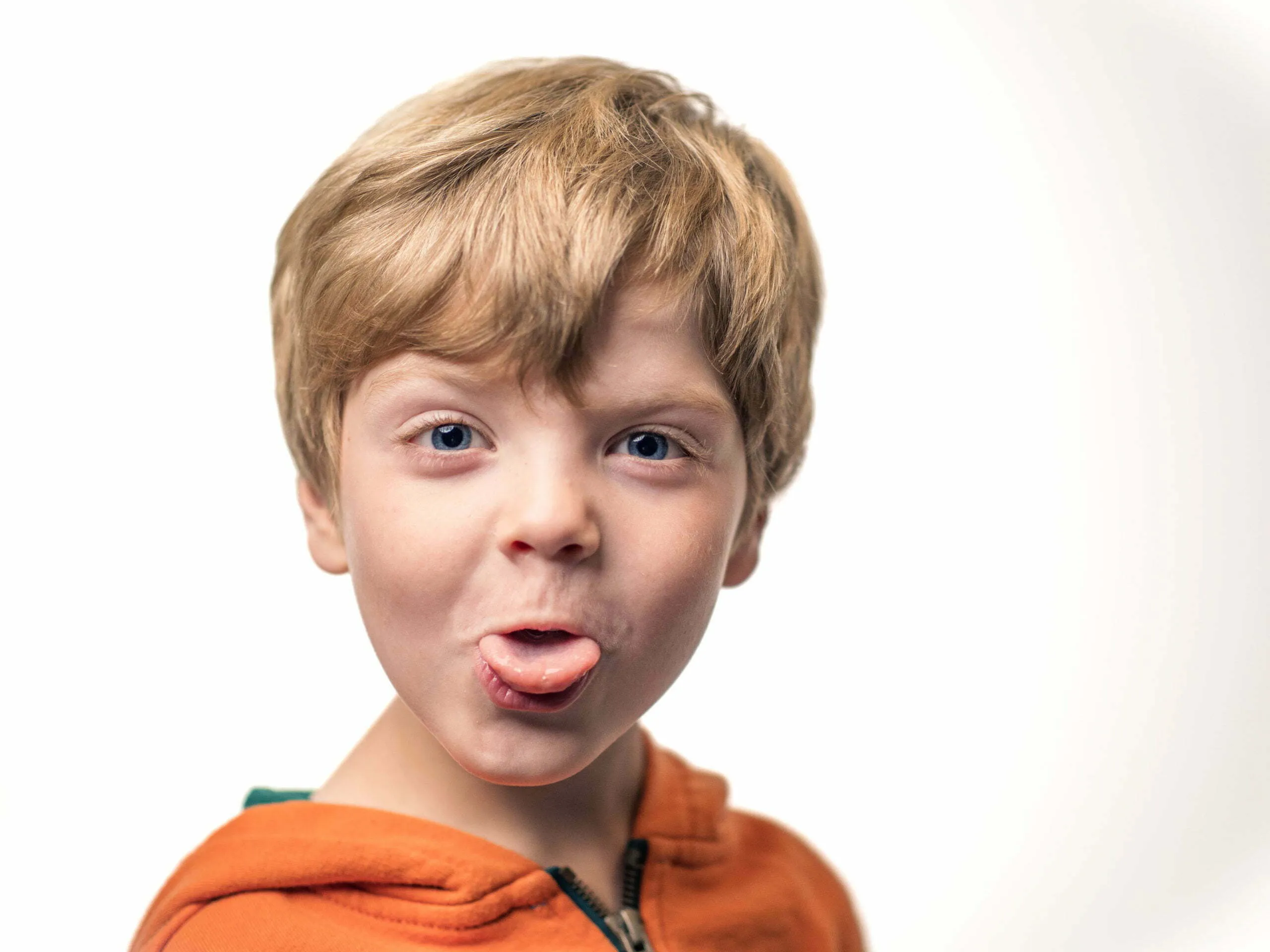 Blonde boy around age seven in orange hoodie sticking out his tongue playfully, white studio background