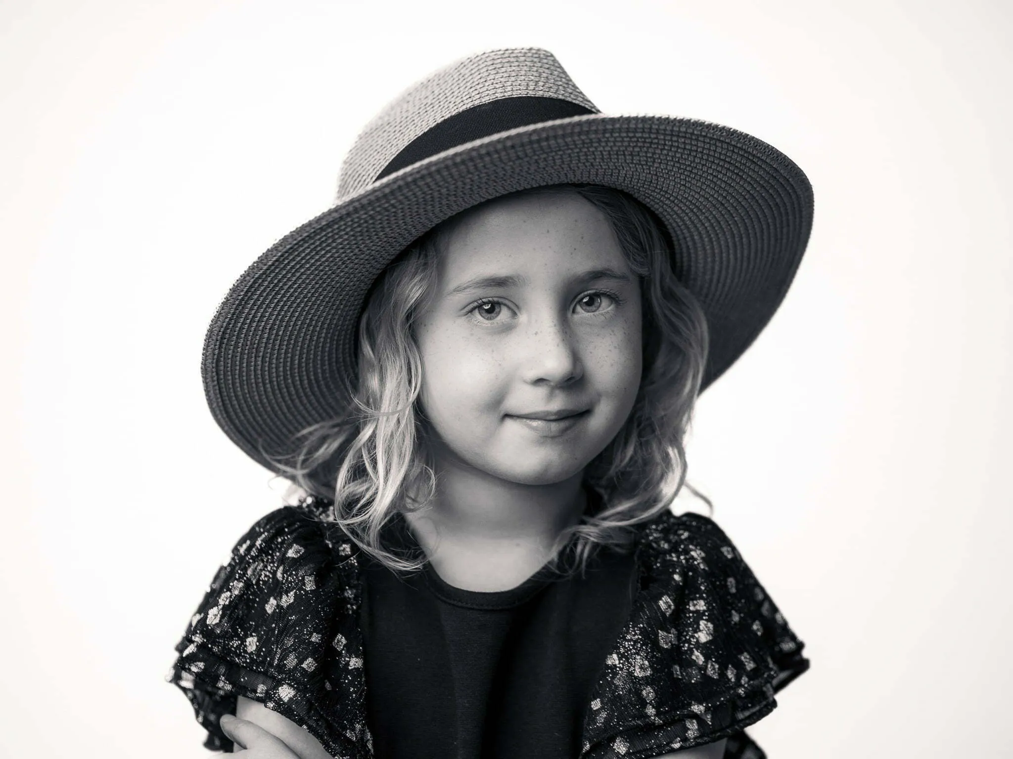 Young girl with curly blonde hair wearing a wide-brim straw hat, smiling softly in black and white studio portrait