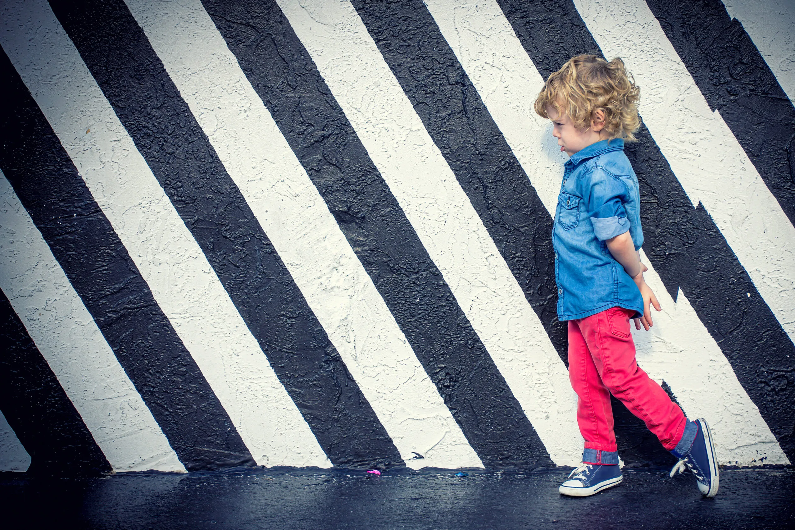 Curly-haired toddler in blue denim shirt and red pants walking past a black and white diagonal striped mural wall