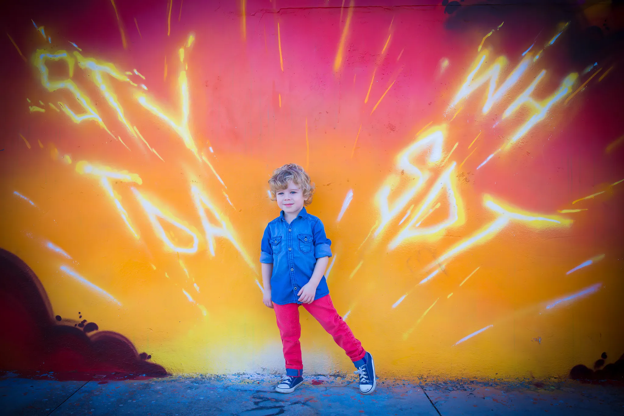 Curly-haired toddler in blue shirt and red pants posing confidently in front of a vibrant pink and orange graffiti mural