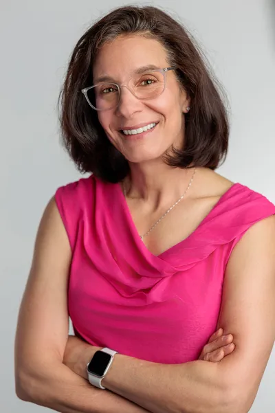 Woman in her 40s wearing a pink blouse and glasses smiling confidently with arms crossed against a light gray studio backdrop