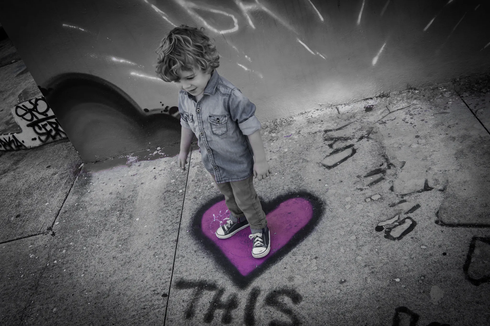 Selective color edit of curly-haired toddler standing on a purple heart painted on a sidewalk near a skate park