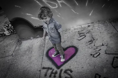 Selective color edit of curly-haired toddler standing on a purple heart painted on a sidewalk near a skate park