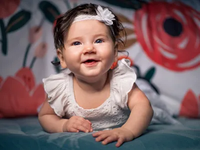 Smiling baby girl with blue eyes and white headband lying on a teal blanket with floral backdrop