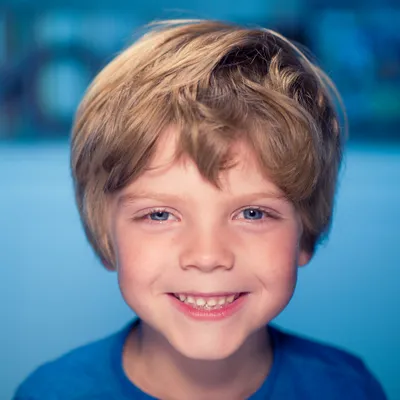 Grinning boy around age seven with tousled blond hair and blue eyes against a soft blue background