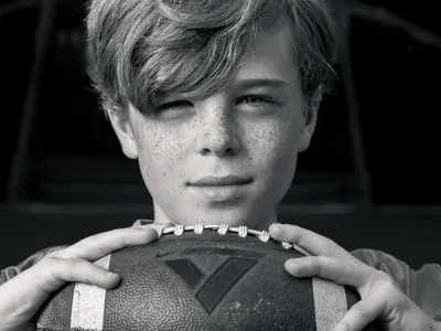 Black and white portrait of a freckled teenage boy resting his chin on a football, direct eye contact