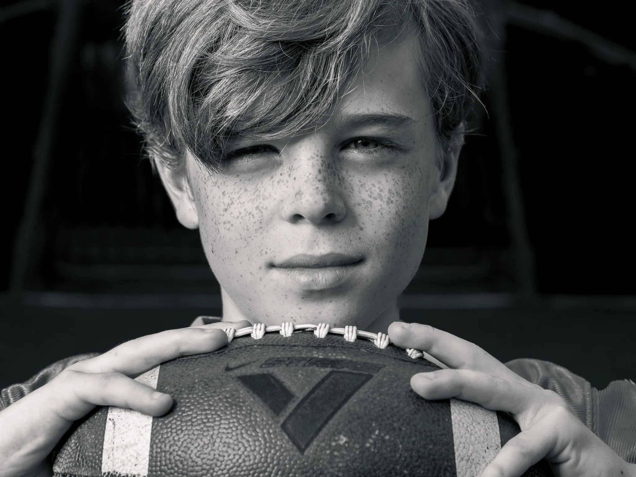Black and white portrait of a freckled teenage boy resting his chin on a football, direct eye contact