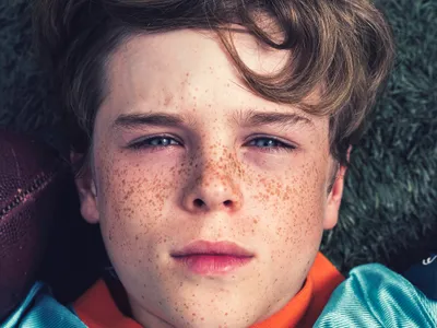 Freckled teen boy lying on grass in a teal and orange jersey with a football beside his head, looking up