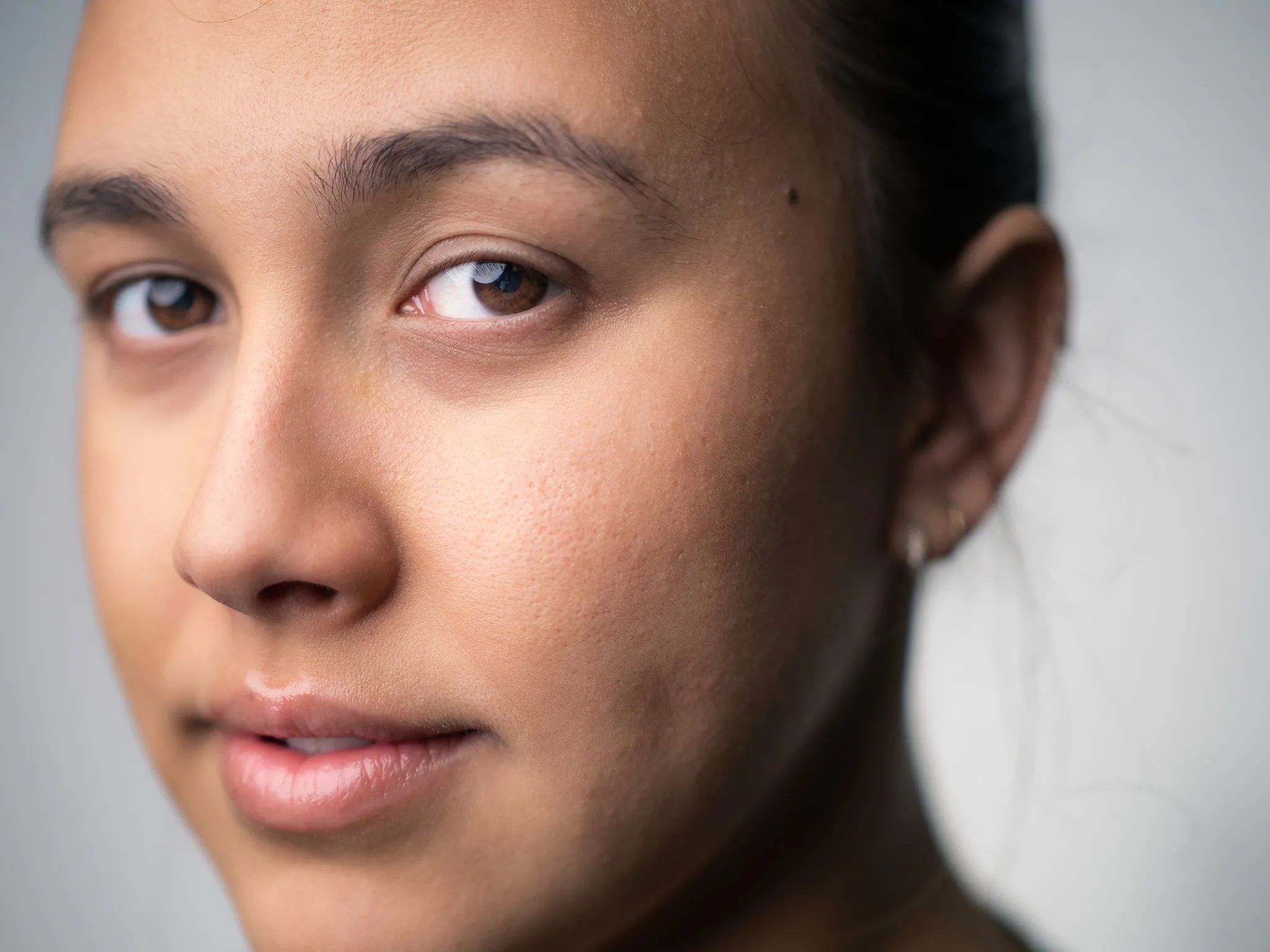 Tight close-up portrait of a teenage girl with brown eyes and pulled-back hair against a gray backdrop