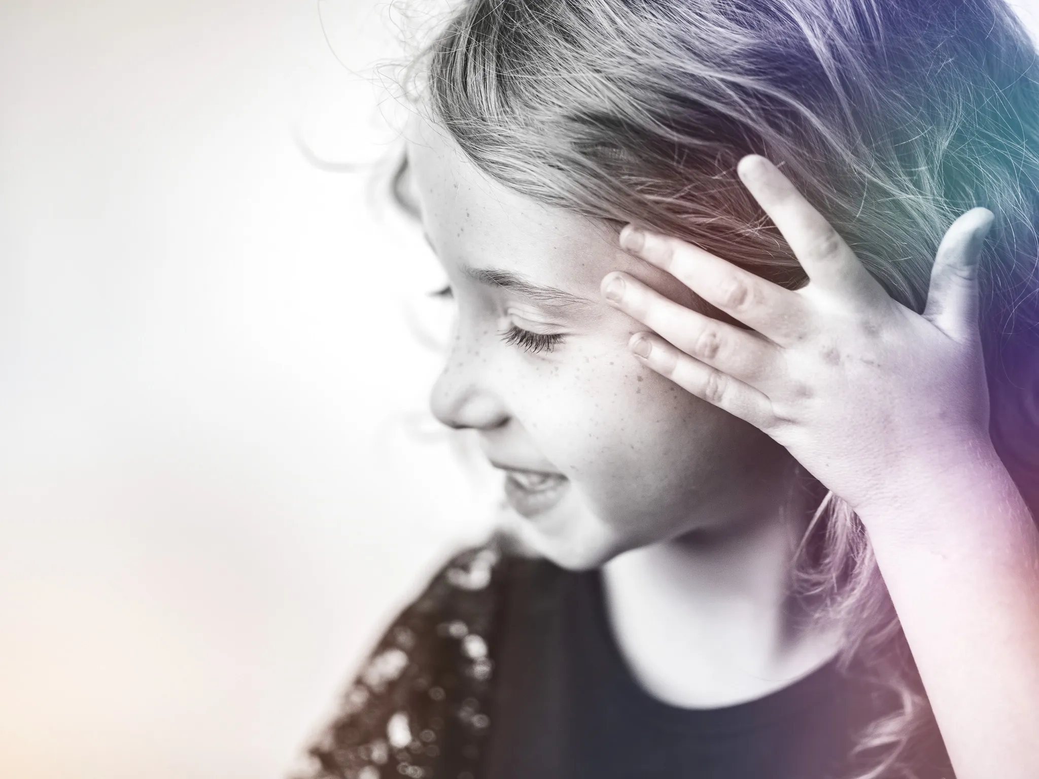 Candid side profile of a young girl laughing with hand brushing hair back, rainbow light in her hair