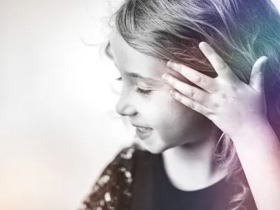 Candid side profile of a young girl laughing with hand brushing hair back, rainbow light in her hair